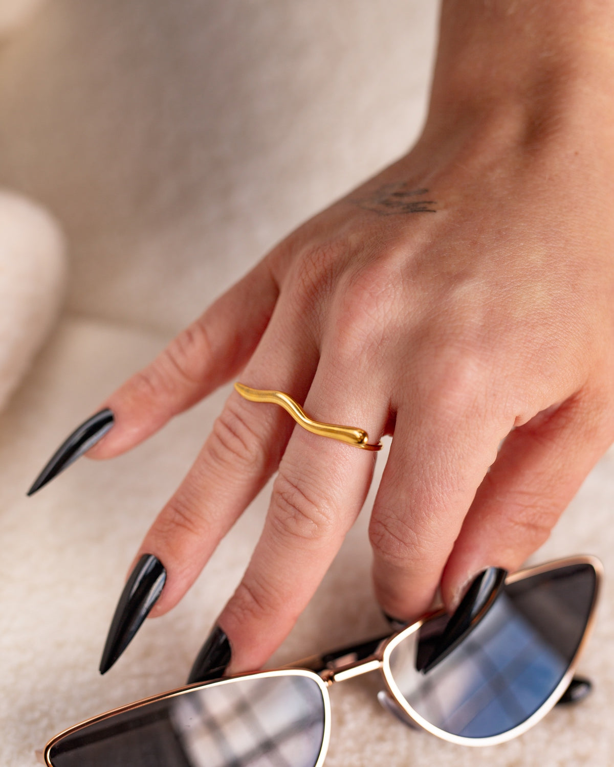 Amelia gold wave bar ring styled with long black nails beside silver sunglasses.