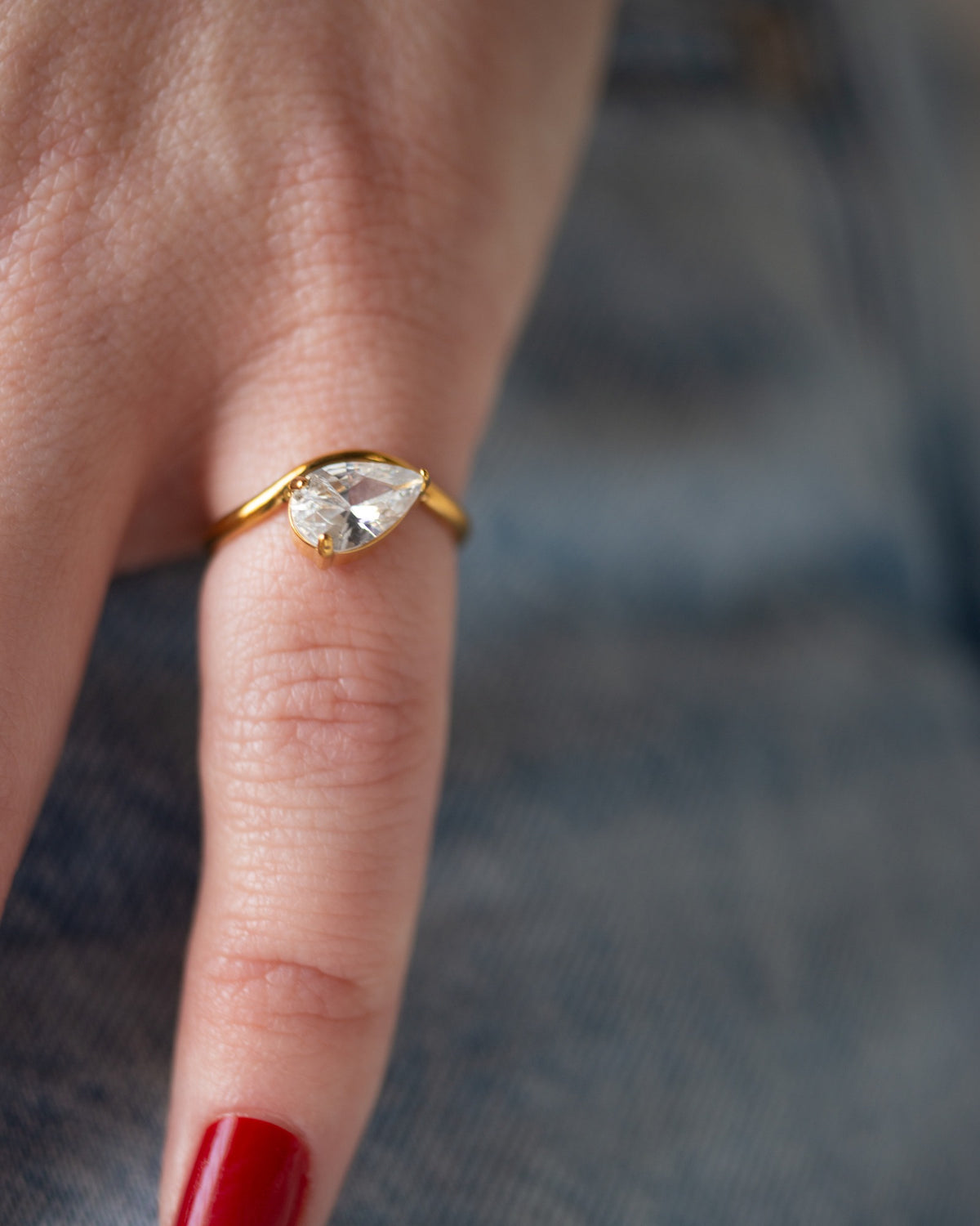 Davina east-west pear-cut ring worn with a long red nail in a tight finger detail against dark denim.