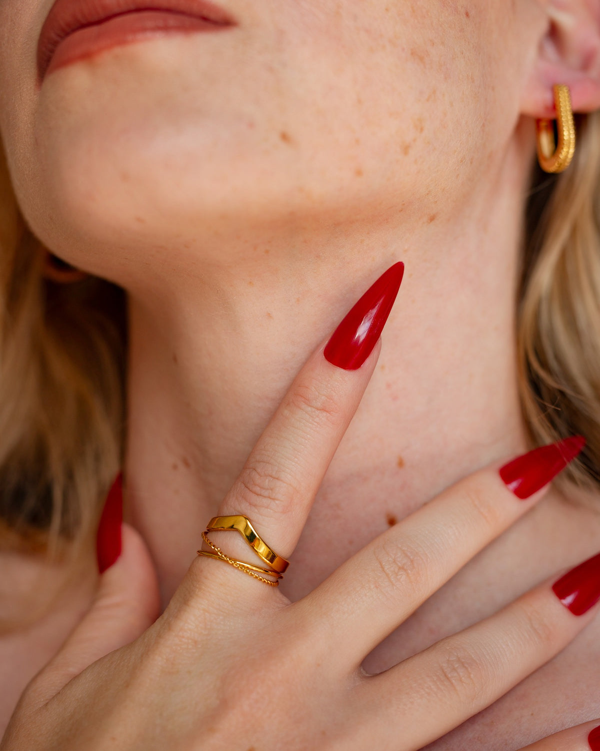 Reina gold chevron stacking ring worn with long red nails in a close hand-to-neck portrait.