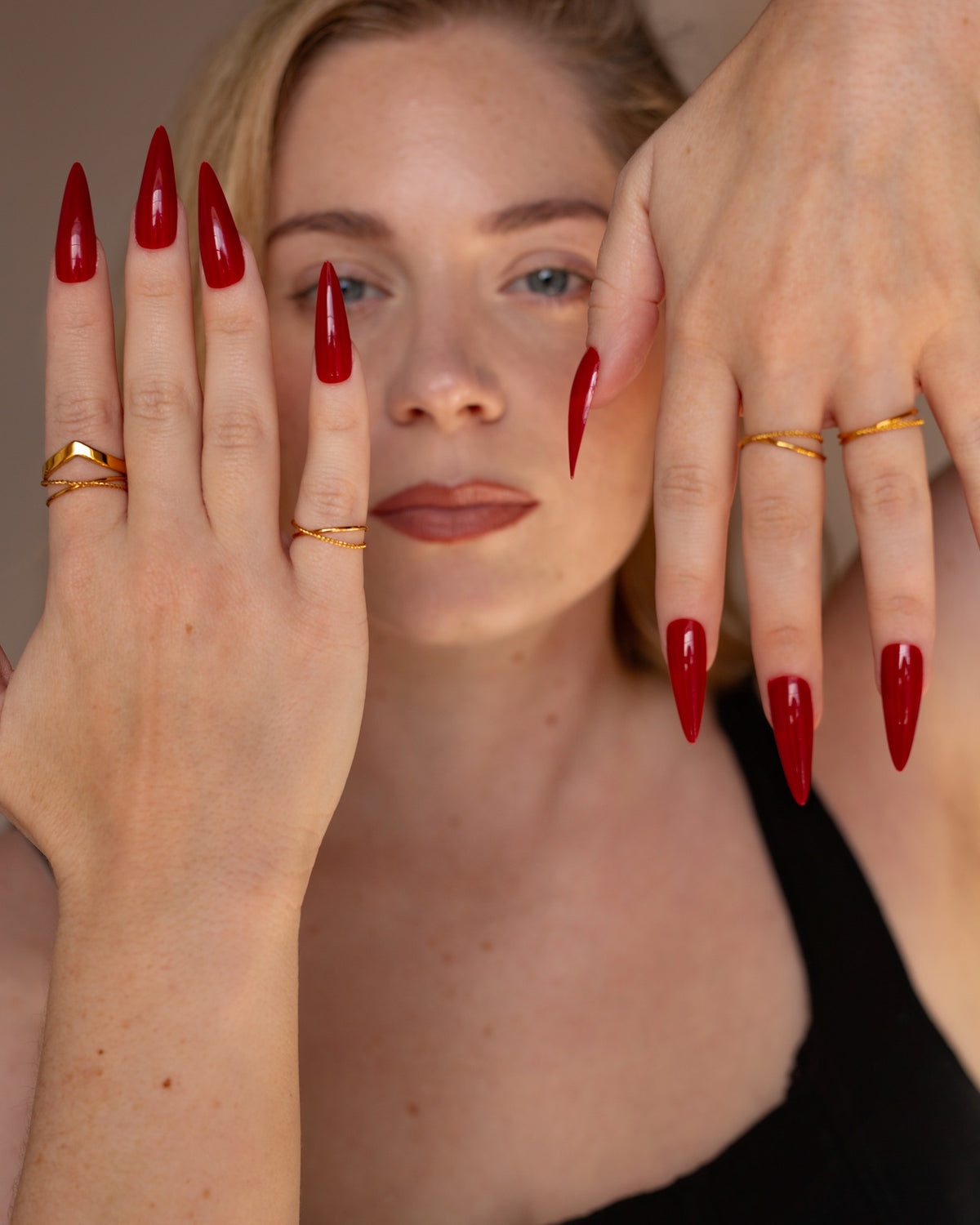 Reina V-shaped gold ring styled with layered rings and long red nails in a two-hand face portrait.