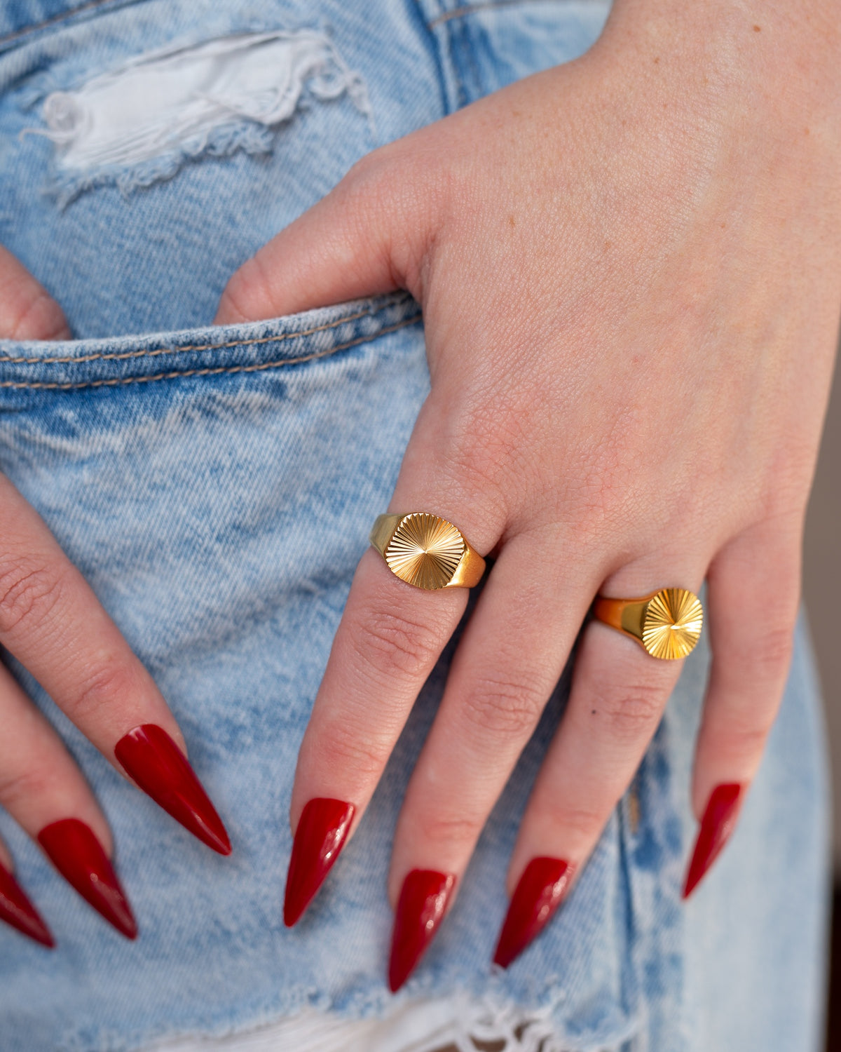 Sol polished gold sunburst signet ring worn with layered rings and long red nails against blue denim.