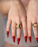 X sculptural crossover ring worn with long red nails in a centered close hand portrait.